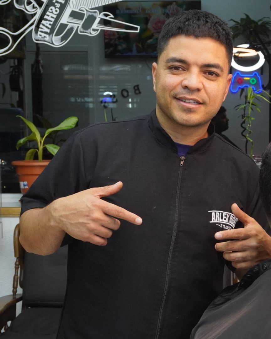 Arlex, con la casaca negra de ARLEX BOSS STUDIOS, señalando
           con el dedo el logotipo bordado en su propio pecho, dentro de su
           barbería