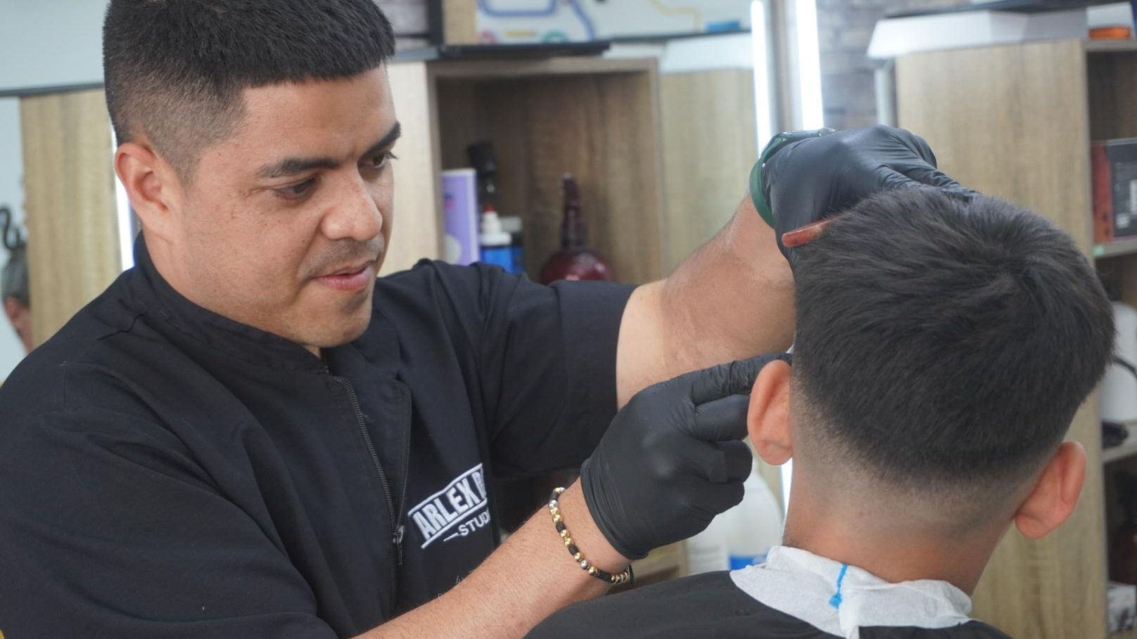 Arlex, con guantes y la casaca de ARLEX BOSS STUDIO, trabajando
         sobre la cabeza de un cliente al que se ve de espaldas, dentro de su
         barbería